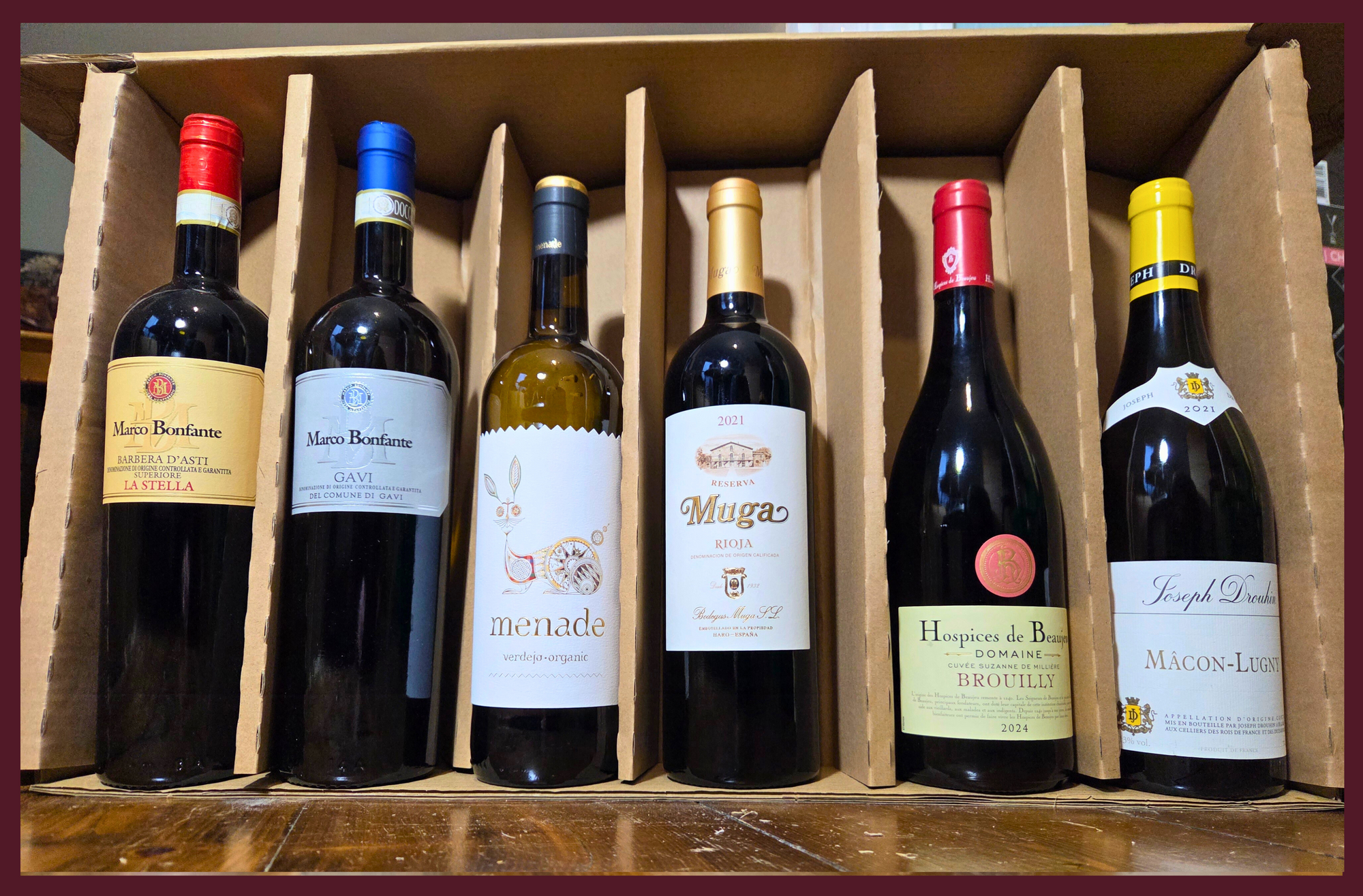 Box of six wine bottles with various labels on a wooden surface.